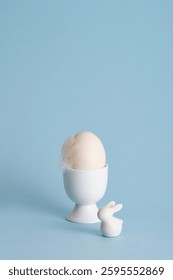 Minimalist Easter egg with a white feather bird and ceramic bunny on a pastel blue background. A place for your text, congratulations, wishes.