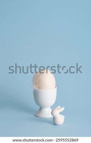Minimalist Easter egg with a white feather bird and ceramic bunny on a pastel blue background. A place for your text, congratulations, wishes.