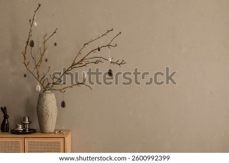 A minimalist and elegant Easter tree with monochromatic hanging decorations, displayed in a sculptural ceramic vase against a textured beige wall.