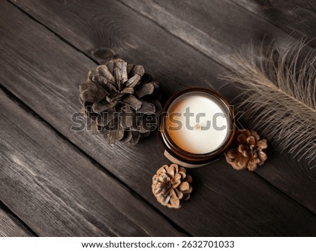 A minimalist flat lay featuring a brown glass candle, pine cones, and a feather on a dark wooden surface. Ideal for seasonal, wellness, or interior design themes. Warm, cozy, and natural atmosphere