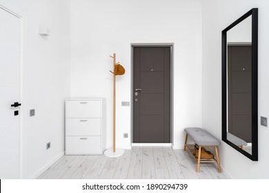 Minimalist hallway with modern interior design in contemporary apartment. Comfortable shoe storage bench near black frame mirror on white wall. Wooden coat hanger in hall with chest drawers - Shutterstock ID 1890294739