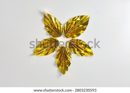A minimalist high angle flat lay photograph showcasing five handmade golden folded paper leaves arranged in a symmetric star pattern, crafted through kid's origami school projects. Isolated on a clean warm white background with negative space, the image represents children's creativity, naive DIY craft, and the highly tactile sensory texture of metallic gold foil.