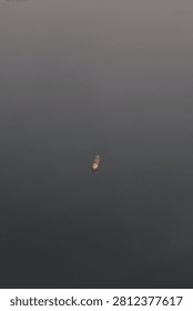 A minimalist, high-angle shot capturing a vast, still body of water shrouded in dense, slate-grey fog or twilight, where the horizon line is completely obscured by the mist. Floating in the exact center of this muted, empty expanse is a solitary boat, its small, warm yellow and orange lights providing a sharp, glowing contrast against the dark surroundings. Along the extreme top edge of the frame, just barely visible, are a few tiny, distant specks of land or faint lights.