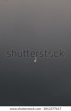 A minimalist, high-angle shot capturing a vast, still body of water shrouded in dense, slate-grey fog or twilight, where the horizon line is completely obscured by the mist. Floating in the exact center of this muted, empty expanse is a solitary boat, its small, warm yellow and orange lights providing a sharp, glowing contrast against the dark surroundings. Along the extreme top edge of the frame, just barely visible, are a few tiny, distant specks of land or faint lights.