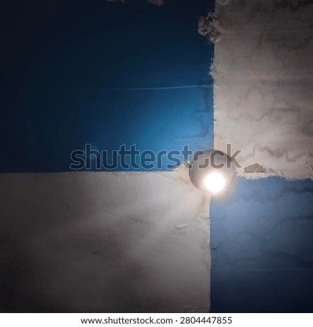 Minimalist interior ceiling with a glowing light bulb against blue and white painted walls. Abstract architectural composition featuring geometric shapes, light, shadow, and textured surfaces in a residential space.