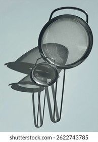 Minimalist kitchen composition featuring two stainless steel mesh strainers casting artistic, overlapping shadows on a light surface. Captured in natural sunlight, the image highlights texture, form