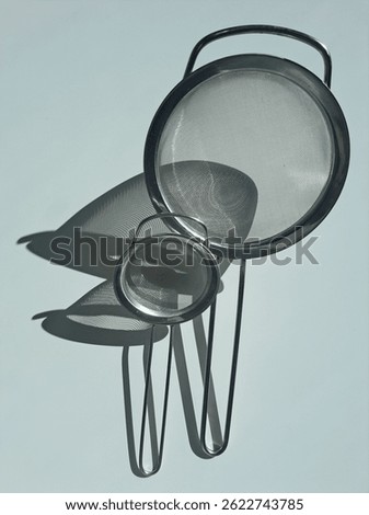 Minimalist kitchen composition featuring two stainless steel mesh strainers casting artistic, overlapping shadows on a light surface. Captured in natural sunlight, the image highlights texture, form