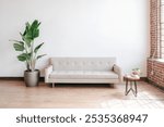 Minimalist living room with a beige sofa, large potted plant, and small round table. Bright, airy space with wooden floors and brick walls. Cozy and inviting.