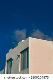 Minimalist low angle architecture of a white concrete building facade with large glass windows under a deep blue sky with soft clouds. 