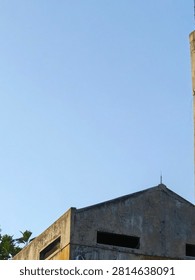 A minimalist, low-angle photograph capturing the angular roofline of an aged, weathered industrial building. The concrete facade shows significant texture, stains, and wear. A small, sharp lightning rod stands prominently at the peak of the gable against a vast, clear, light blue sky. The composition emphasizes geometry, urban decay textures, and negative space. This image is ideal for backgrounds, conceptual design, or themes related to architecture, aging infrastructure, and minimalism.