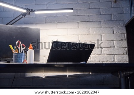 Minimalist office setup featuring closed black laptop, illuminated adjustable desk lamp, and stationery holder with scissors, glue, and pens on dark desk in front of white painted brick wall