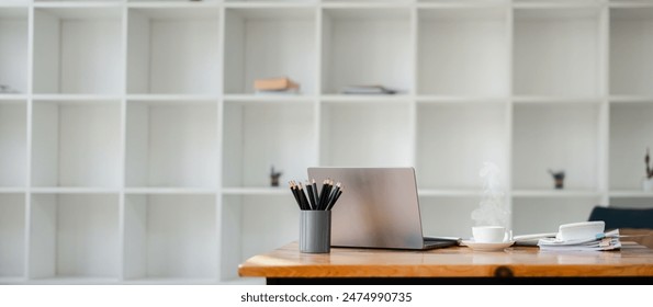 Minimalist office setup featuring a laptop, coffee cup, and stationery on a wooden desk with white shelves in the background. - Powered by Shutterstock