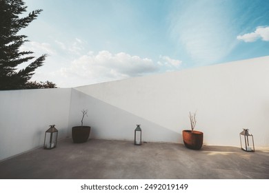 Minimalist outdoor terrace with a white wall, three clay pots containing small plants, and two decorative lanterns. The sky is clear with a few clouds, creating a serene and clean atmosphere - Powered by Shutterstock