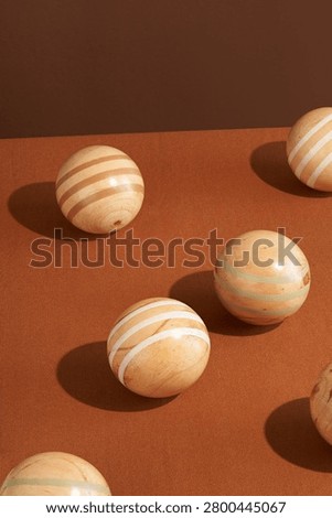 A minimalist studio shot featuring several smooth spherical pétanque natural wooden balls arranged on a solid textile terracotta brown surface with defined shadows. Abstract arrangement of natural wooden balls on a warm brown tabletop. Rounded shapes placed in a simple still life composition with open negative space. Smooth wood texture and light beige tones visible on the surface of the balls. Strong directional light creating soft elongated shadows. Minimal Scandinavian styling with geometric forms and earthy colors. Decorative homeware photograph with natural materials and clean balance. Quiet tabletop scene focused on shape, texture and composition.