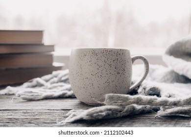 Minimalist Style. Coffee, Cozy Grey Sweater And Books On Vintage Windowsill Against Snow Landscape From Outside. Soft Focus. Relaxing Winter Day At Home With Traditional Winter Hot Drink.