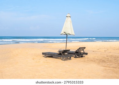 Minimalist view of an empty sandy beach with two dark wooden lounge chairs and a closed sun umbrella. Serene tropical ocean coast with calm waves and blue sky in the background, perfect for vacation and relaxation concept.