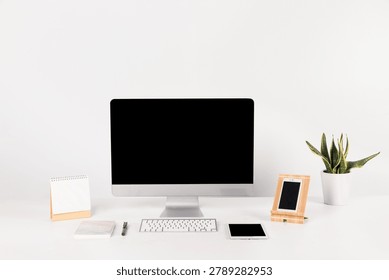 Minimalist workspace featuring a desktop computer monitor with wireless keyboard, tablet, smartphone, notebook, and potted succulent plant on a clean white desk.