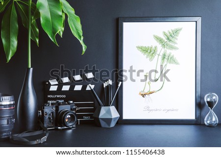 Minimalistic black desk with mock up photo frame, tropical leaf, photo camera, black accessories and plant.s  Black backgrounds wall. Black decor interior.