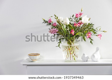 A minimalistic bouquet of white tulips, pink eustoma, hyacinth, eucalyptus in a fluted glass vase on a white panel of an artificial fireplace. Cup of tea, faience figurines of a two birds