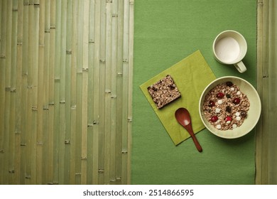 A minimalistic breakfast setting with a bowl of granola topped with dried fruits and white chocolate, a granola bar, and a cup of milk, placed on a green placemat with a wooden spoon. - Powered by Shutterstock