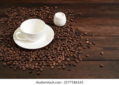 A minimalistic composition featuring an empty white coffee cup on a saucer, accompanied by a small white creamer and surrounded by scattered roasted coffee beans on a rustic dark wooden table.