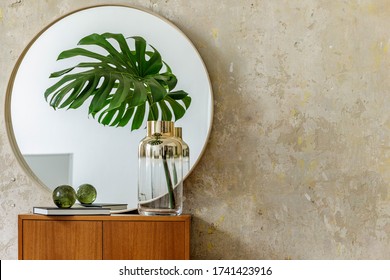 Minimalistic composition at modern retro interior with design commode, tropical leaves in vase, round mirror, copy space, book, grunge wall and elegant accessories in stylish home decor.