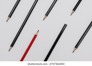 Minimalistic flat lay composition featuring single red colored pencil placed among several black graphite pencils on clean white background. The constrasting color creates strong visual metaphor for individuality, uniqueness, leadership, difference and standing out from the crowd.