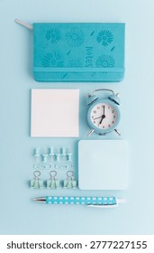 Minimalistic flat lay composition featuring blue stationery items, including a notebook, alarm clock, sticky notes, pen, and clips, arranged on a light blue background. 
