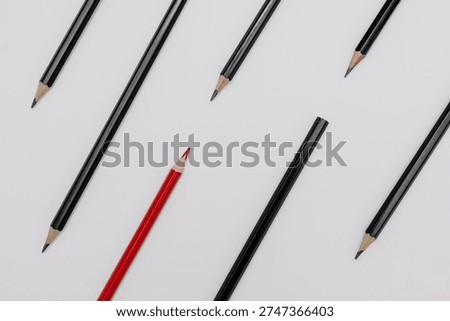 Minimalistic flat lay composition featuring single red colored pencil placed among several black graphite pencils on clean white background. The constrasting color creates strong visual metaphor for individuality, uniqueness, leadership, difference and standing out from the crowd.