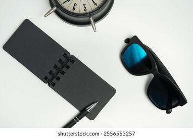 A minimalistic flat lay featuring a black notebook, pen, sunglasses, and vintage alarm clock placed on a white background, perfect for themes of organization and style. - Powered by Shutterstock