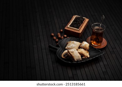 Minimalistic scene with traditional pastries on a black plate, hazelnuts, and a glass of tea. A rustic wooden box with loose tea leaves enhances the cozy, authentic tea-time atmosphere. - Powered by Shutterstock