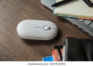 A minimalistic workspace with a white wireless computer mouse, notebook, pen, laptop, and books on a wooden desk. Perfect for technology and remote work concepts.