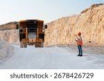 Mining engineer holding a tablet while directing a dumper truck in an open pit mine, overseeing mineral extraction operations in a dynamic landscape