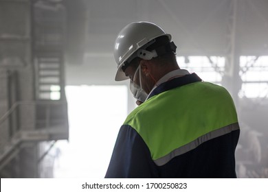 Mining Engineer In Yellow-blue Uniform And White Helmet And Respirator Inspects Dusty And Dirty Mining Workshop