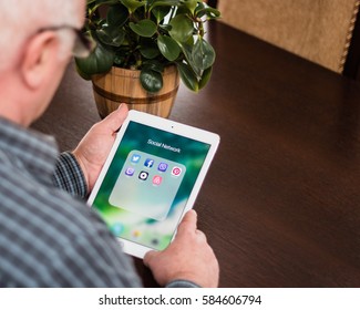 Minsk, Belarus - February 18, 2017: Old Gray-haired Man Pensioner Is Sitting Near The Desk, Holding IPad Air Apple. Social Networks Icons. Twitter. Facebook. Skype. Viber. Pinterest. Uber. Airbnb.