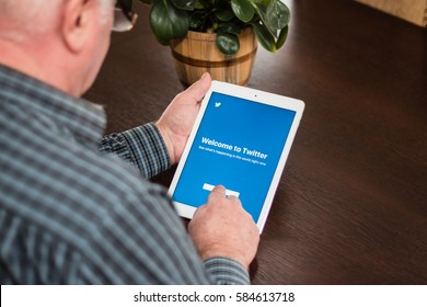 Minsk, Belarus - February 18, 2017: Old Gray-haired Man Pensioner Sits Near The Desk, Hold IPad Air Apple.  He Is Trying To Create An Account On The Popular Social Network Twitter.