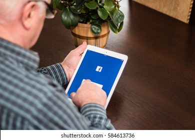 Minsk, Belarus - February 18, 2017: Old Gray-haired Man Pensioner Sits Near The Desk, Hold IPad Air Apple.  He Is Trying To Create An Account On The Popular Social Network Facebook.