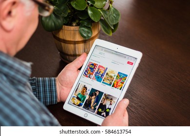 Minsk, Belarus - February 18, 2017: Old Gray-haired Man Pensioner Sits Near The Desk, Hold IPad Air Apple.  He Read About Airbnb And Look Its Page On The Web Site In Internet.