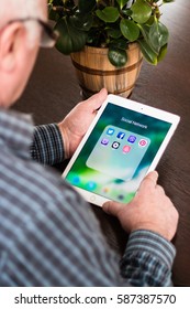 Minsk, Belarus - February 18, 2017: Old Gray-haired Man Pensioner Is Sitting Near The Desk, Holding IPad Air Apple. Social Networks Icons. Twitter. Facebook. Skype. Viber. Pinterest. Uber. Airbnb.