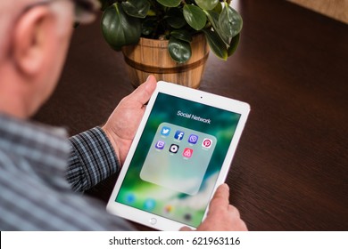 Minsk, Belarus - February 18, 2017: Old Gray-haired Man Pensioner Is Sitting Near The Desk, Holding IPad Air Apple. Social Networks Icons. Twitter. Facebook. Skype. Viber. Pinterest. Uber. Airbnb.