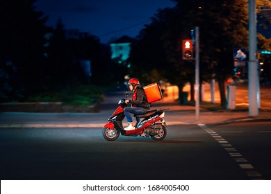 Minsk, Belarus. Jul 2019. Delivery Man Delivering Online Food Orders On Moto Scooter At Night. Young Delivery Boy With Red Thermal Bag, Restaurant Courier Carries Food, Quick Deliver Meal To Costumer