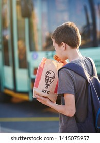 Minsk, Belarus - May 2019. Hungry Kid Eats Kentucky Fried Chicken At Bus Stop. Teenager Eating Bare Hands KFC Hot Wings From Paper Bag Pack At Street. Boy Eating Junk Fast Food At City Street