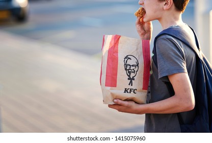 Minsk, Belarus - May 2019. Hungry Kid Eats Kentucky Fried Chicken At Bus Stop. Teenager Eat With Dirty Hands KFC Hot Wings From Paper Bag At Street. Boy Eating Junk Fast Food At City Street