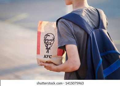 Minsk, Belarus - May 2019. Hungry kid eats Kentucky Fried Chicken at bus stop. Teenager with backpack hold in hands paper bag with food from KFC at street. Boy eating fast food at city street