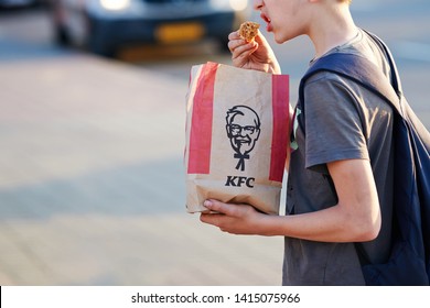 Minsk, Belarus - May 2019. Hungry Kid Eats Kentucky Fried Chicken At Bus Stop. Teenager With Backpack Eat Bare Hands KFC Hot Wings From Paper Bag At Street. Boy Eating Junk Fast Food At City Street