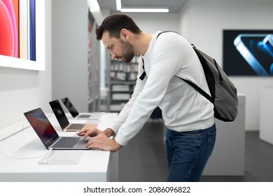 Minsk, Belarus - Nov 27, 2021: Photo Of Young Handsome Thinking Brunet Man With Stubble In A White Sweatshirt Chooses A Computer Laptop For Work And Studying In An Electronic Store In A Shopping Mall