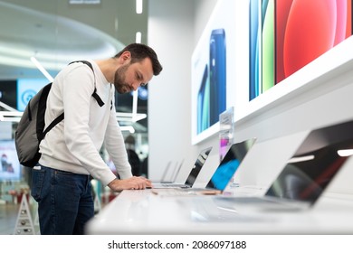 Minsk, Belarus - Nov 27, 2021: Photo Of An Adult Handsome Thinking Brunet Man With Stubble In A White Sweatshirt Chooses A Computer Laptop For Work And Studying In An Electronic Store In A Shopping