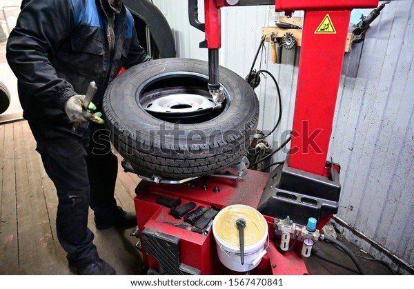 Minsk, Belarus - November 19,\
2019: Photography with a tire fitting. A worker is serving a car.\
He puts the tire on the wheel disc using a special machine and\
tool.