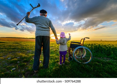 Miracle Recovery: Old Man Gets Up From Wheelchair And Raises Hands Up. Overjoyed Grandfather Standing Up And Holding Hand Of His Granddaughter. Shot In A Meadow. Recovery Concept