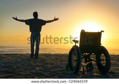 Miracle spiritual healing man with a disability exalted arms spread at the ocean shoreline as he stands up out of his wheelchair and walks towards the sunrise